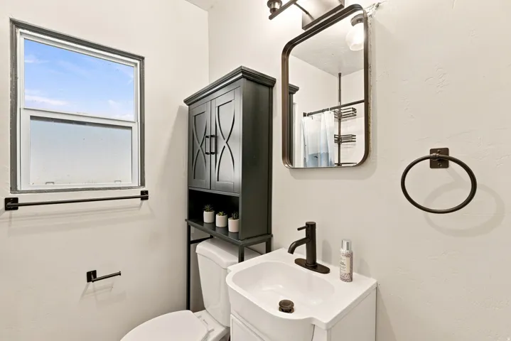 White sink basin with a matte black faucet, framed mirror with integrated lighting, window with dark trim, and a black storage cabinet