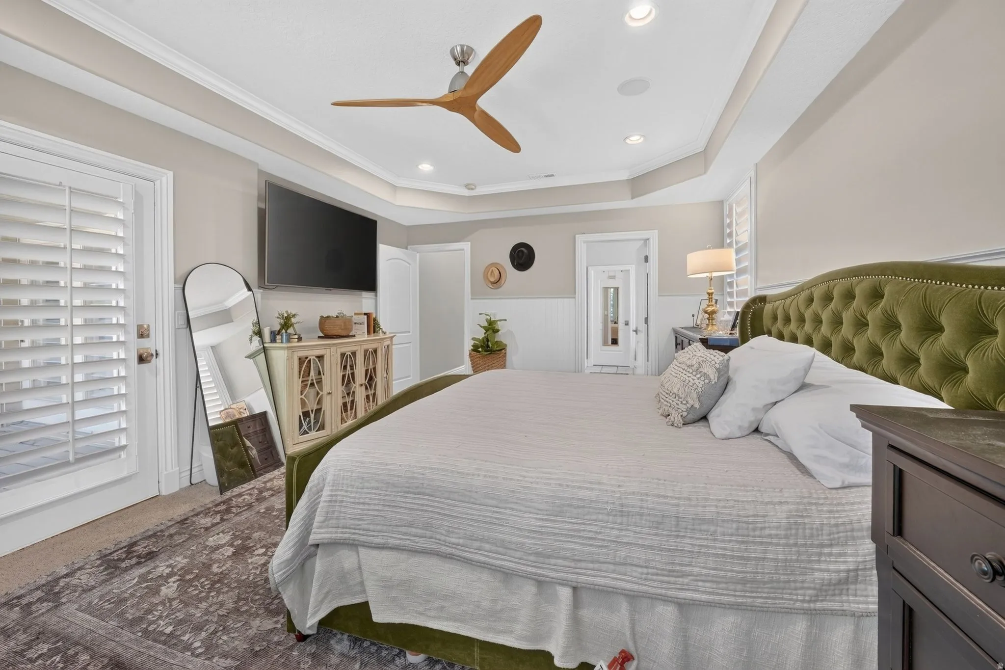 Tray ceiling with recessed lighting, crown molding, wood-blade ceiling fan, white plantation shutters, and wainscoting