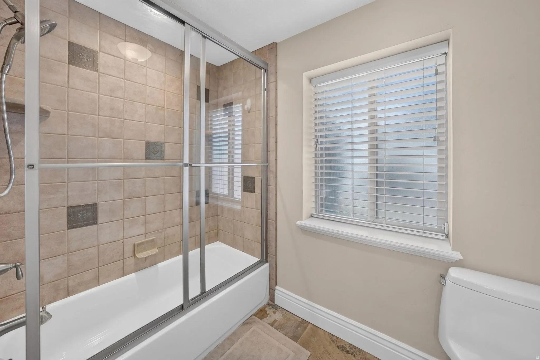 Tub and shower combination with sliding glass doors, neutral tile surround with decorative inlays, and window with privacy blinds