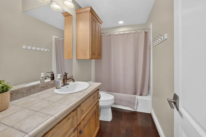 Built-in wood-finish cabinetry, tile countertop, undermount sink with dark bronze fixtures, overhead wood-finish storage cabinet, and wood-finish flooring