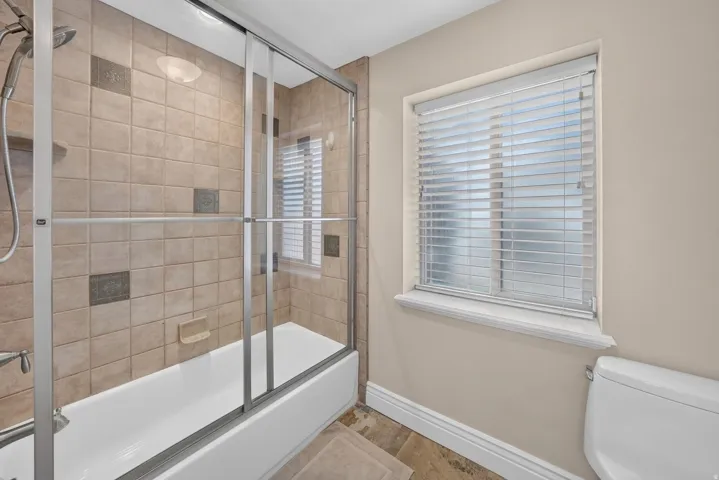 Tub and shower combination with sliding glass doors, neutral tile surround with decorative inlays, and window with privacy blinds