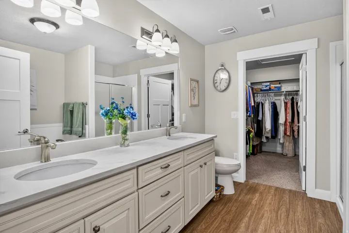 Bathroom featuring a dual vanity with white countertops, wood-finish flooring, a full-width mirror, and an adjacent walk-in closet
