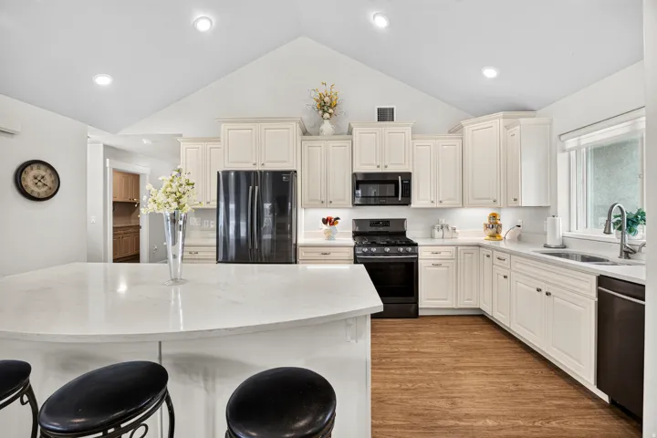 Vaulted ceiling kitchen featuring white cabinetry, black appliances, wood-finish flooring, and a curved island with a light-toned countertop