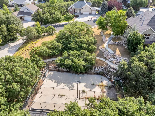 Fenced concrete court surrounded by mature trees, featuring a paved pathway with rock borders and terraced landscaping