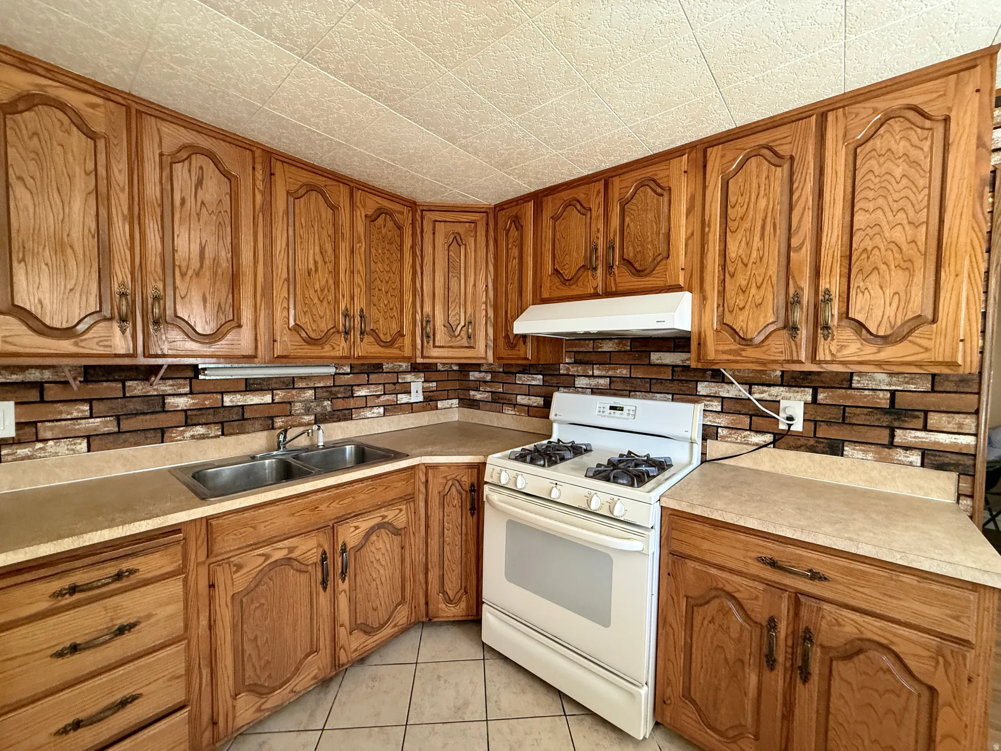 Wood-finish cabinetry with decorative panels, brick-patterned backsplash, light-toned countertops, and a white gas range