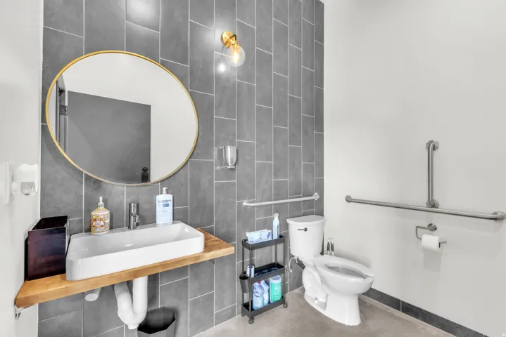 Modern bathroom featuring a rectangular vessel sink on a wood-finish floating vanity, a gold-framed circular mirror, and gray vertically-stacked tile