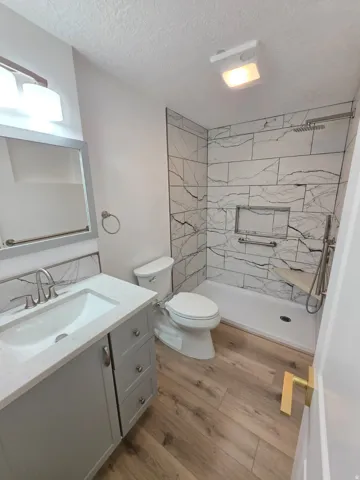 Wood-finish flooring, gray vanity with white countertop, chrome faucet, white subway tile shower surround with black veining, and a built-in shower niche