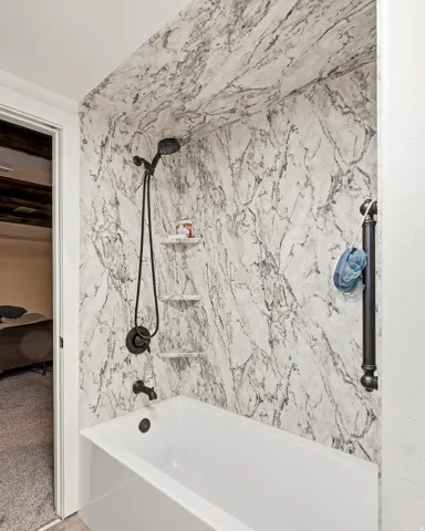 White bathtub with matte black fixtures, marble-patterned shower surround, and built-in corner shelves
