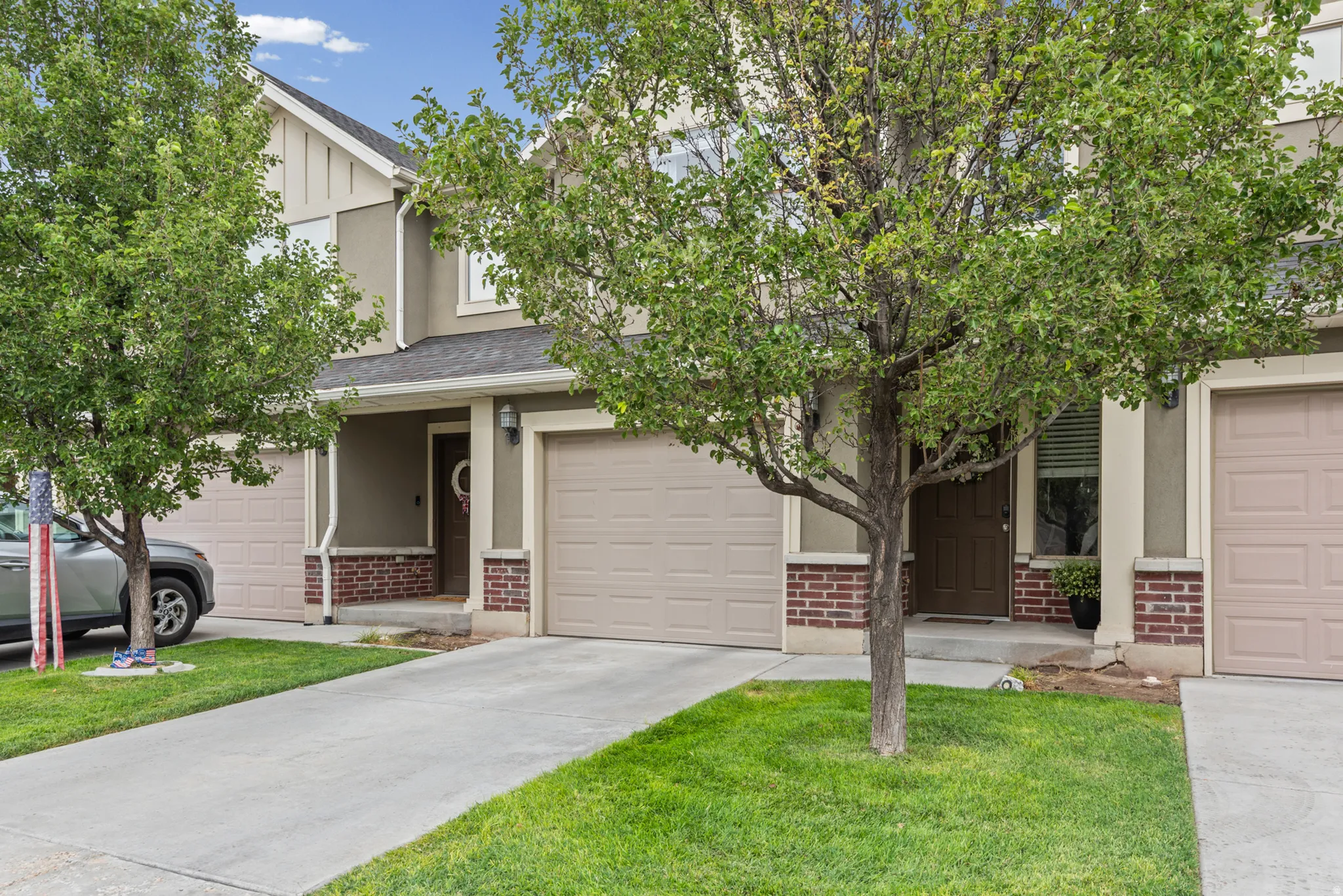 Two-story townhome featuring a stucco and brick facade, attached garages, mature trees, and a concrete driveway