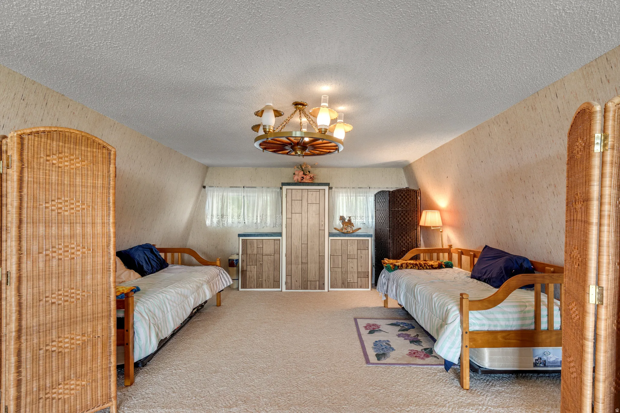Spacious secondary bedroom suite featuring two twin wood-frame beds with coordinating bedding, woven rattan room dividers, carpet flooring, ample natural light from multiple windows, and a ceiling-mounted fixture with decorative brass accents