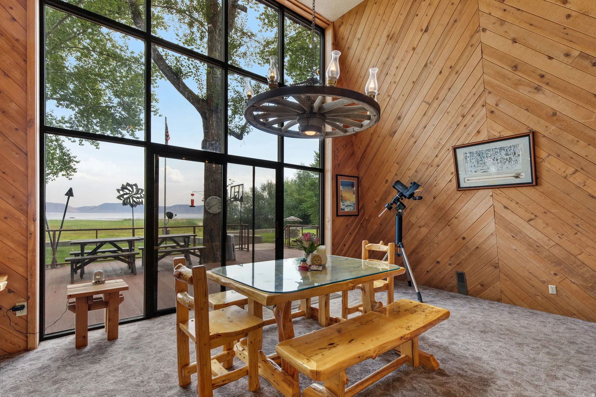 Expansive floor-to-ceiling windows, wood-paneled accent wall, exposed wood beam ceiling, contemporary wagon wheel chandelier, and durable carpet flooring