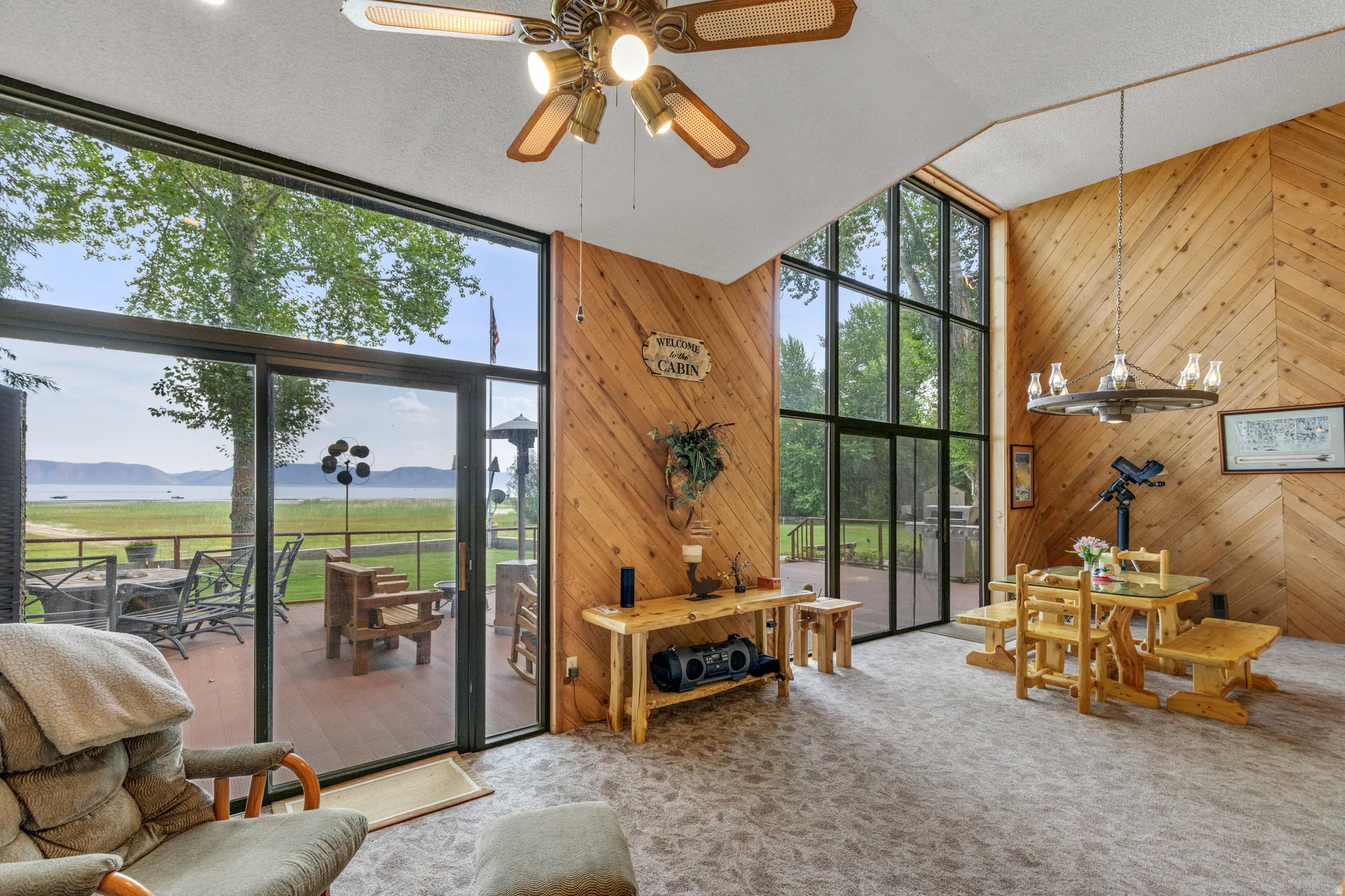 Spacious living area showcasing dramatic wood-finish herringbone accent wall, large multi-panel glass doors and windows framing expansive lawn and landscape vistas, polished concrete flooring, and a brass ceiling fan with integrated lighting