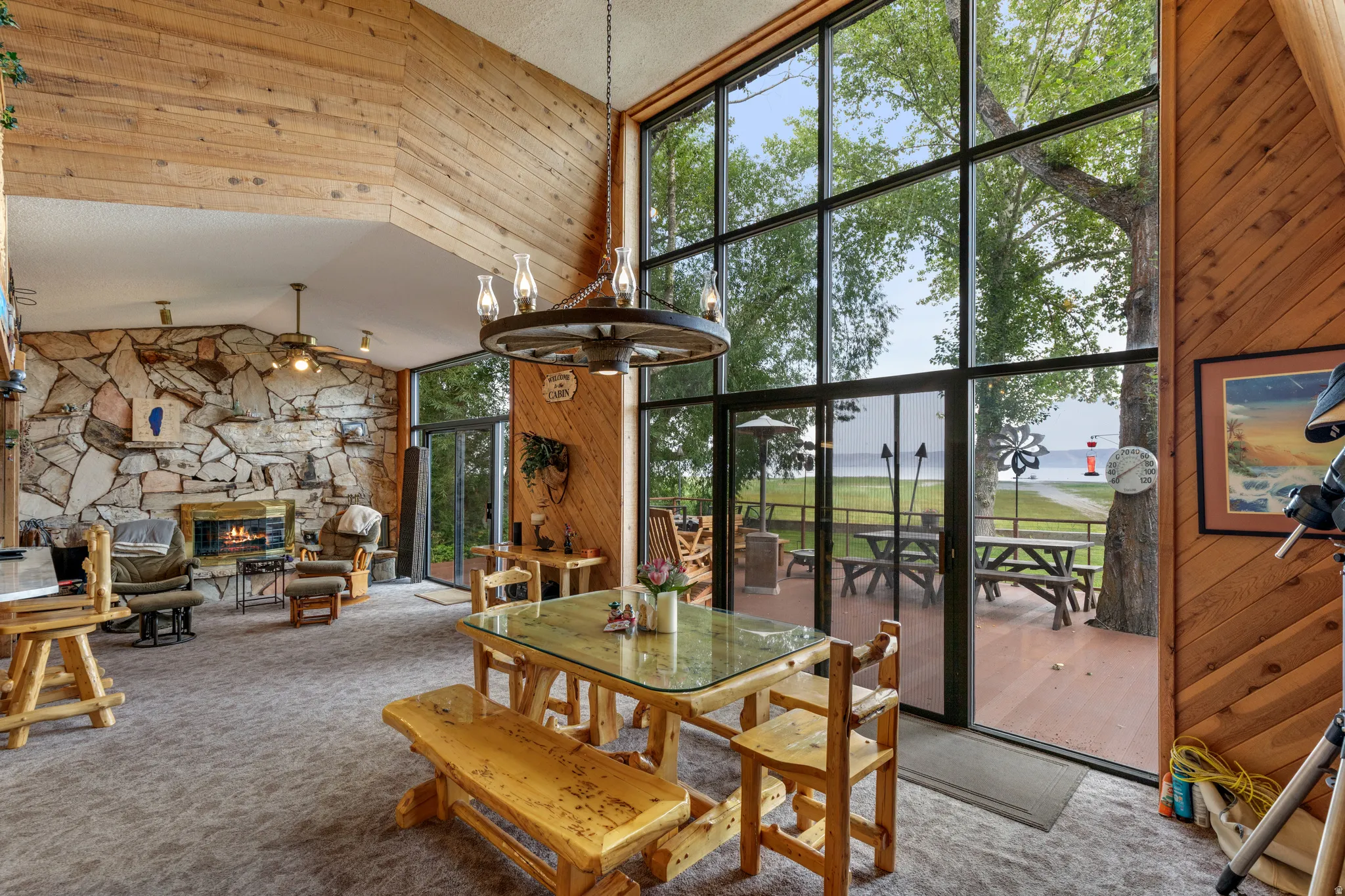Expansive window wall, stone-clad fireplace, wood-finish walls, deck access, and overhead lighting fixture