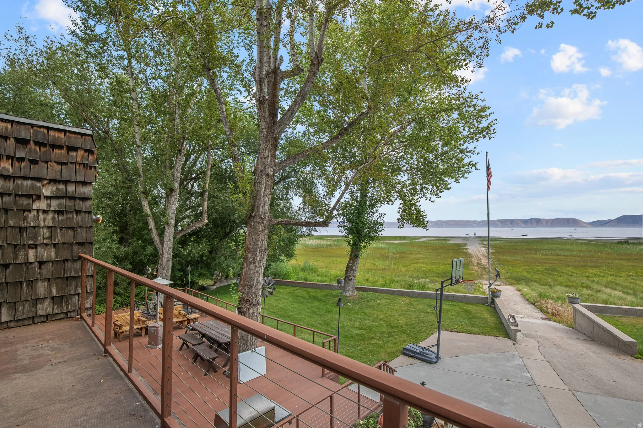 Expansive waterfront property featuring a large composite deck, shingle siding, mature trees, and a concrete pathway to the water