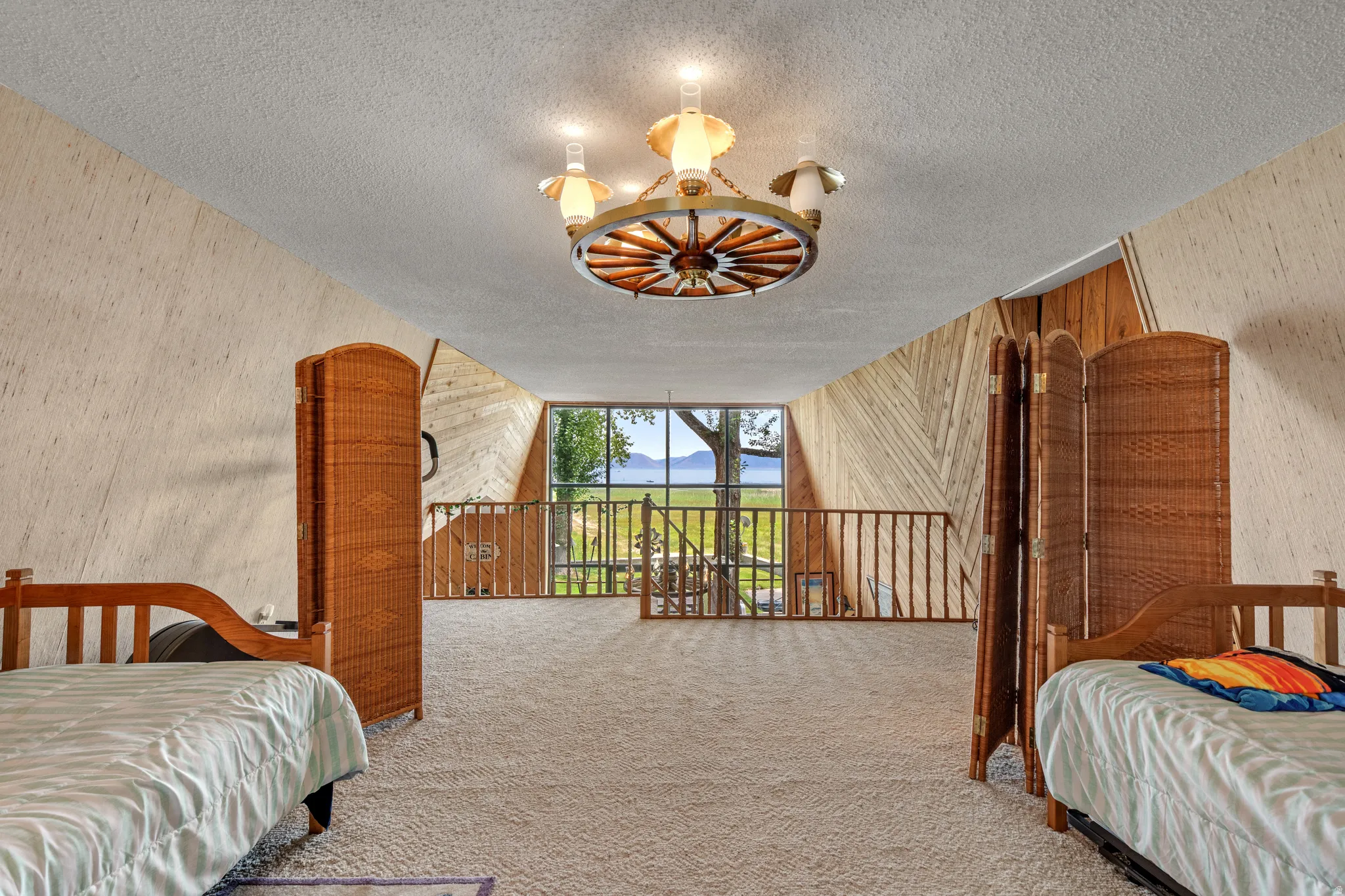 Vaulted ceiling with wagon wheel light fixture, carpeted flooring, and a railing overlooking an exterior landscape