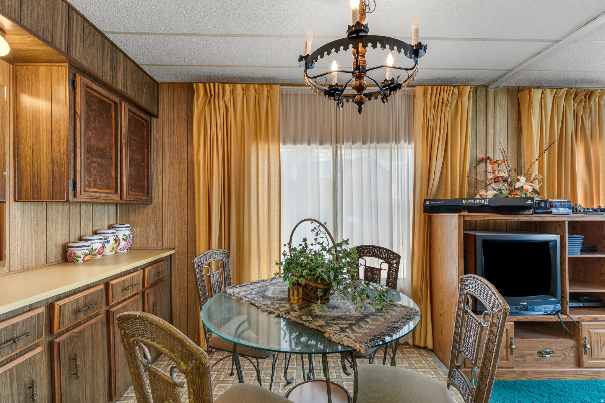 Wood-paneled walls and cabinetry, recessed ceiling with central chandelier, and large windows with privacy sheers