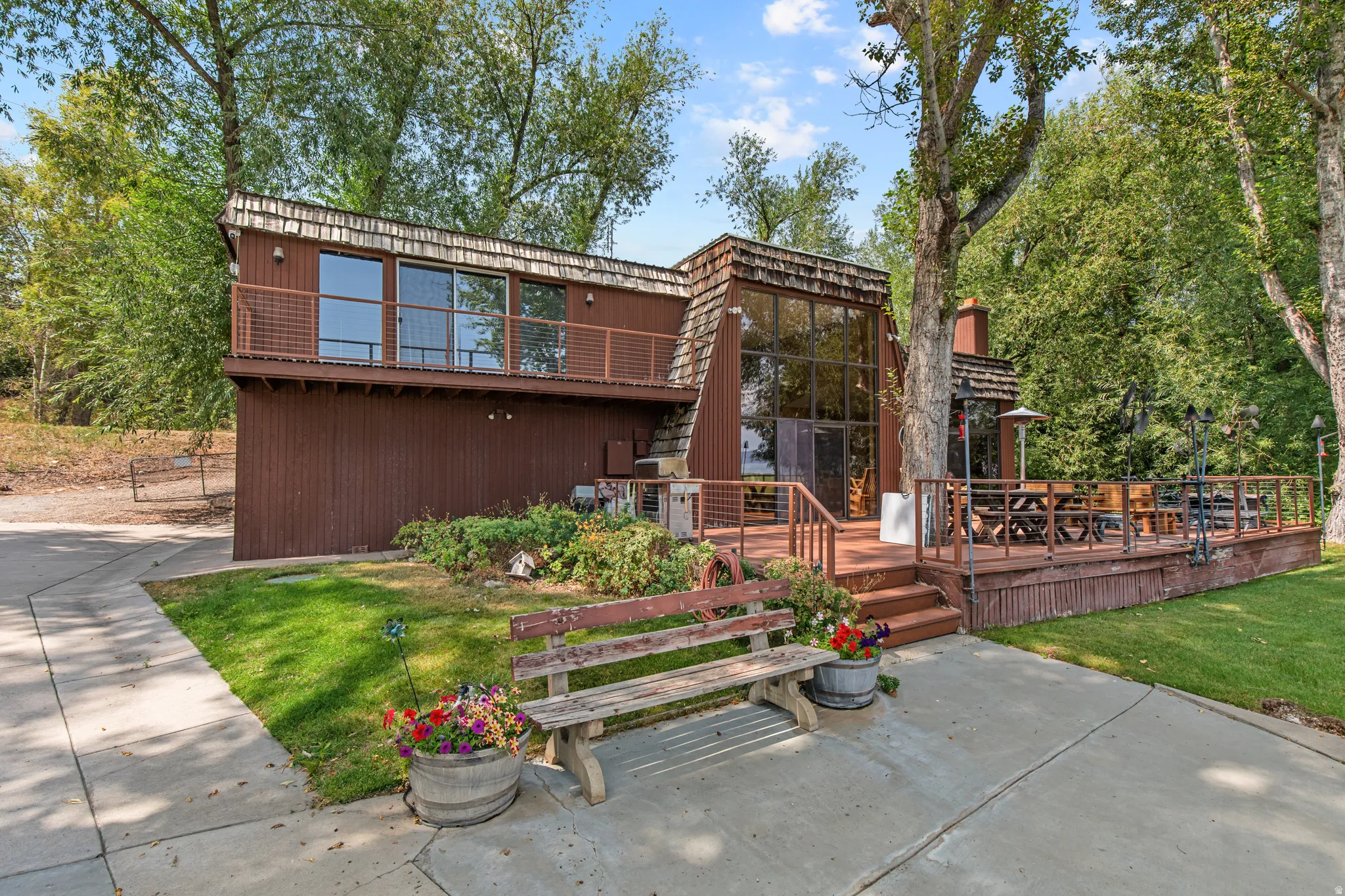 Multi-level brown-sided exterior with a large wood deck, a second-story balcony with metal railings, an A-frame wall of windows, and a concrete pathway