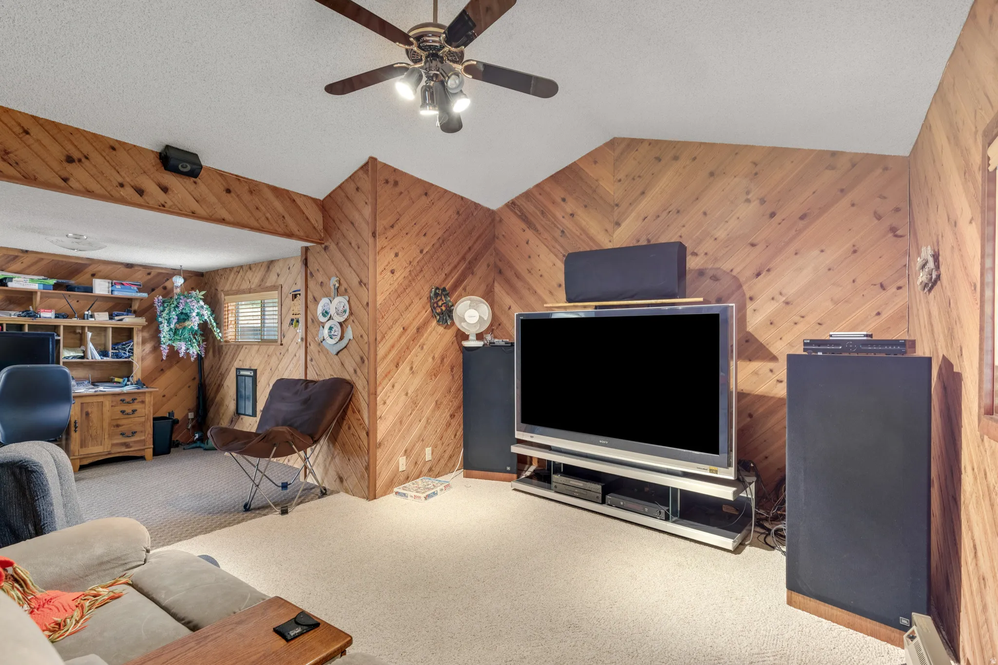 Wood-paneled accent wall with diagonal pattern, textured ceiling, ceiling fan with integrated lighting, window with blinds, and built-in shelving in an adjacent area
