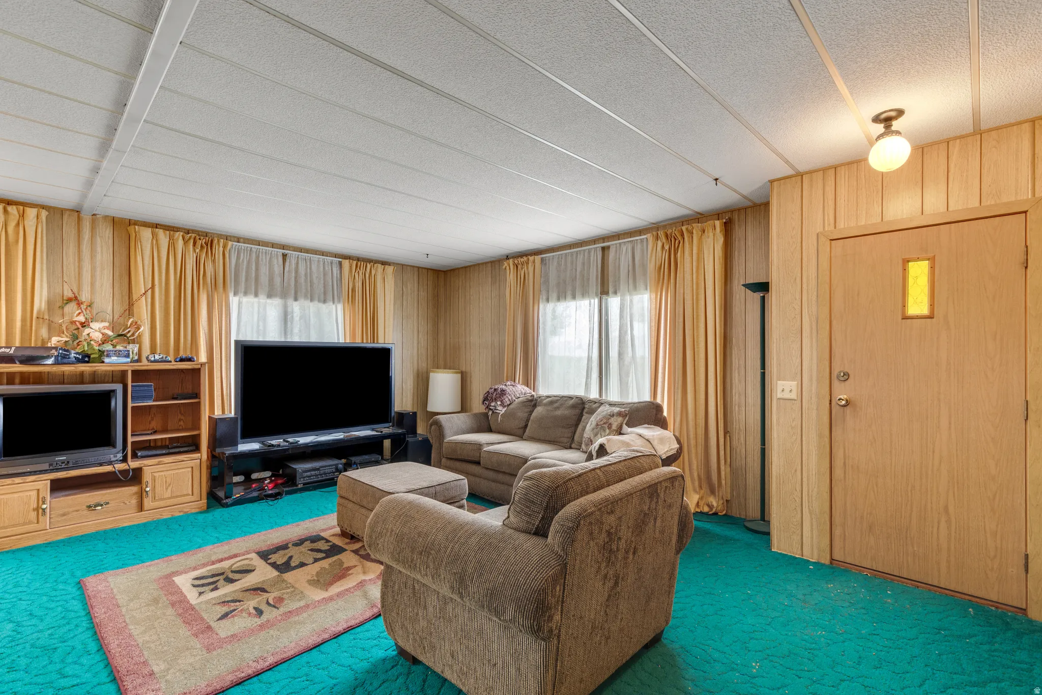 Spacious recreation room featuring wood-paneled walls, a white drop ceiling with recessed lighting, built-in wood cabinetry with media storage, and a vibrant turquoise carpet