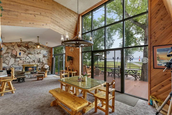 Expansive window wall, stone-clad fireplace, wood-finish walls, deck access, and overhead lighting fixture