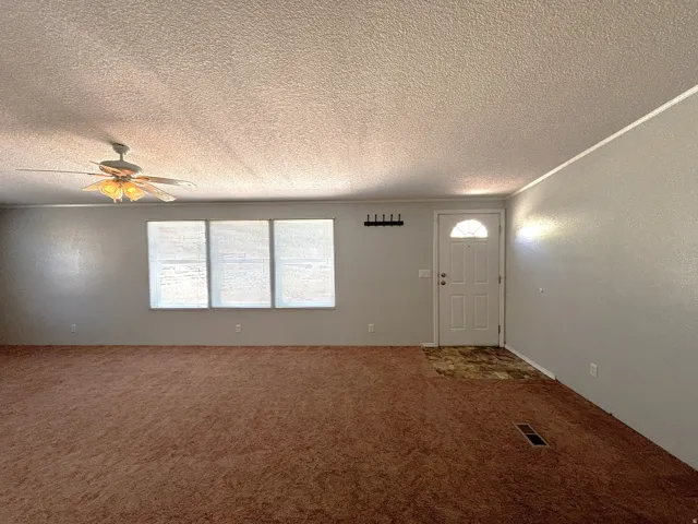 Spacious room featuring carpeting, multiple windows, a ceiling fan with integrated lighting, and a paneled entry door with an arched window
