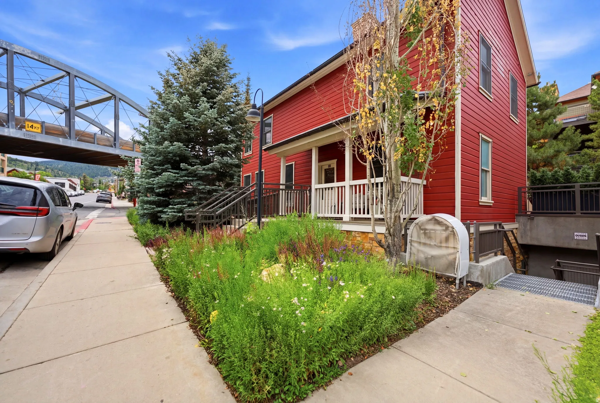 Red exterior siding with a covered front porch, concrete sidewalk, street-level landscaping, and a metal pedestrian bridge overhead