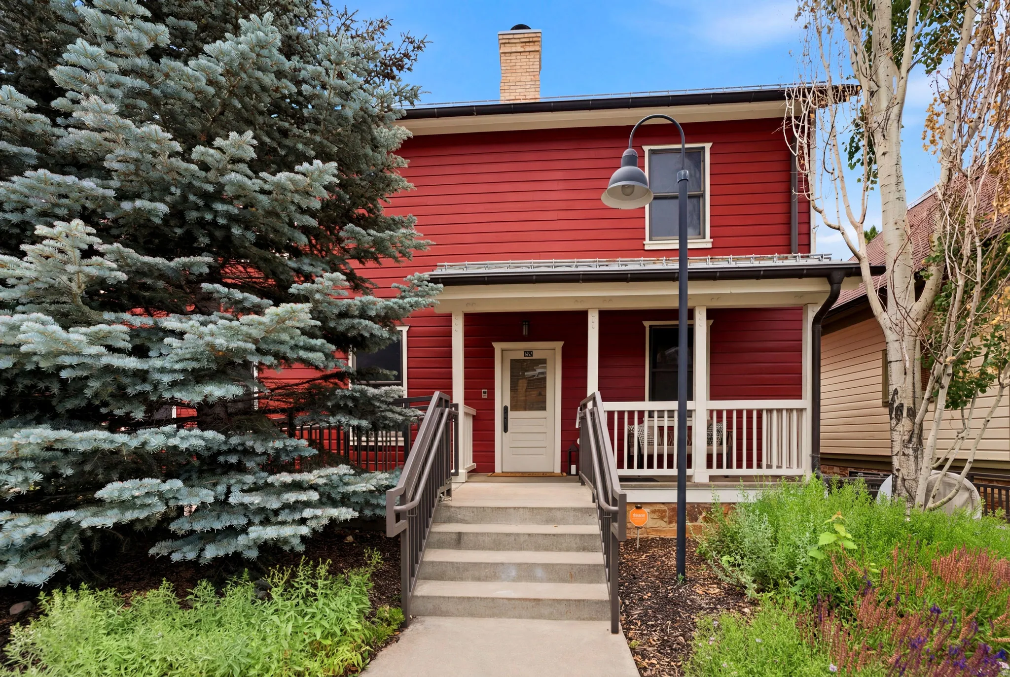 Red exterior siding, inviting front porch, white door with decorative glass, concrete entry steps with handrails, and a mature evergreen tree