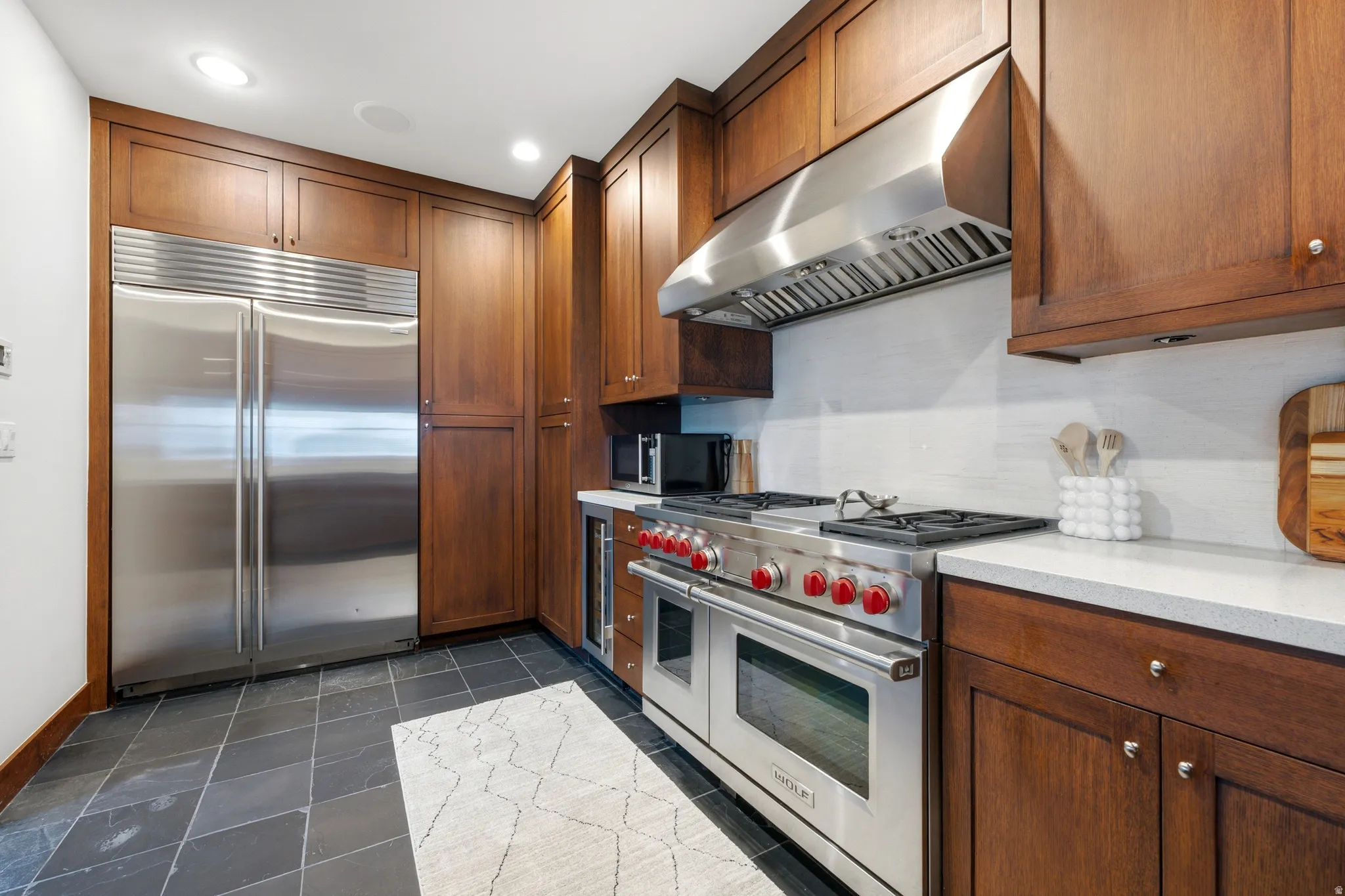Stainless steel refrigerator, built-in range with hood, dark wood cabinetry, light stone countertops, and dark tile flooring