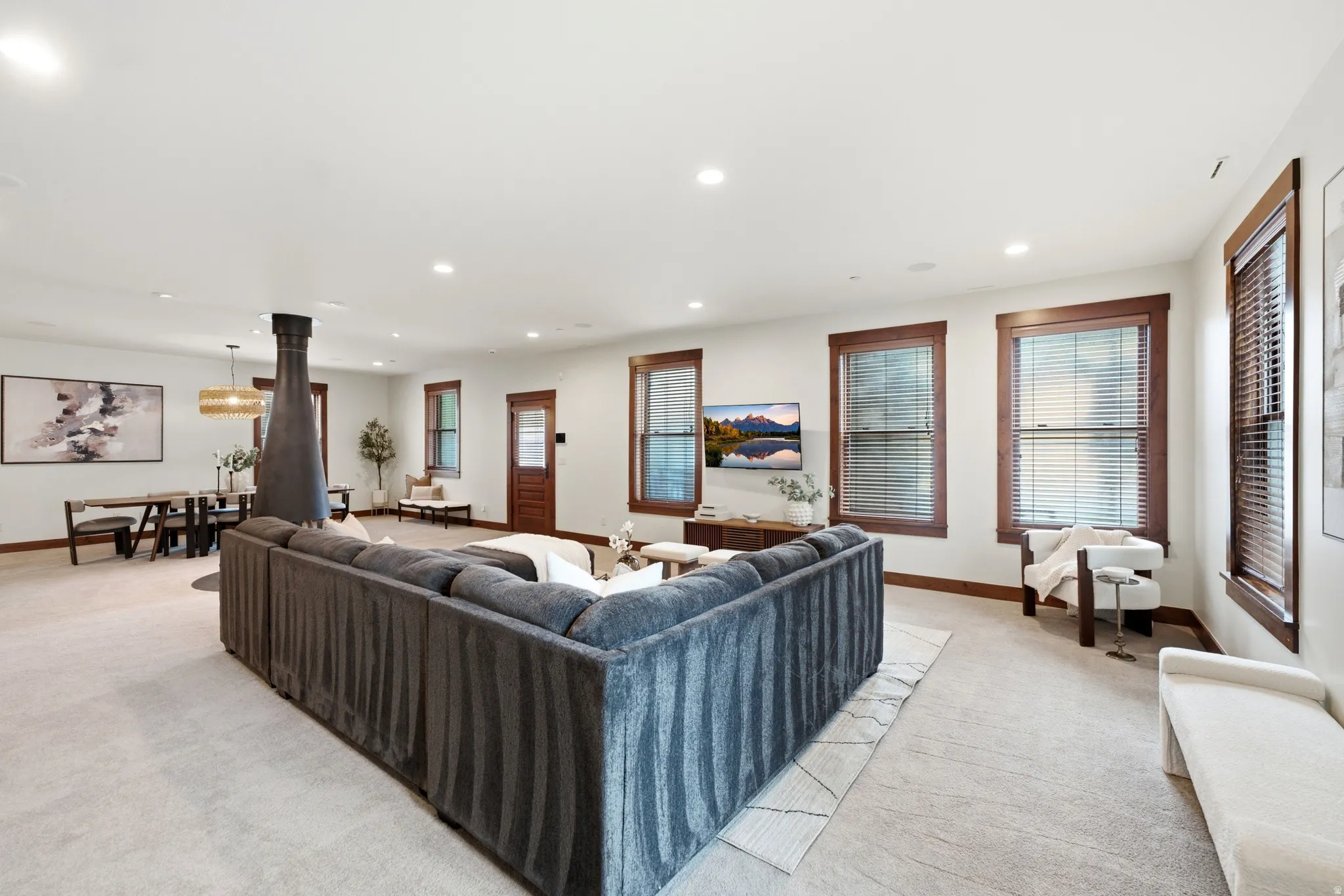 Expansive great room anchored by a striking central fireplace with black metal surround, featuring a curved sectional sofa arrangement, recessed ceiling lighting, and abundant natural light from multiple sets of French doors and transom windows