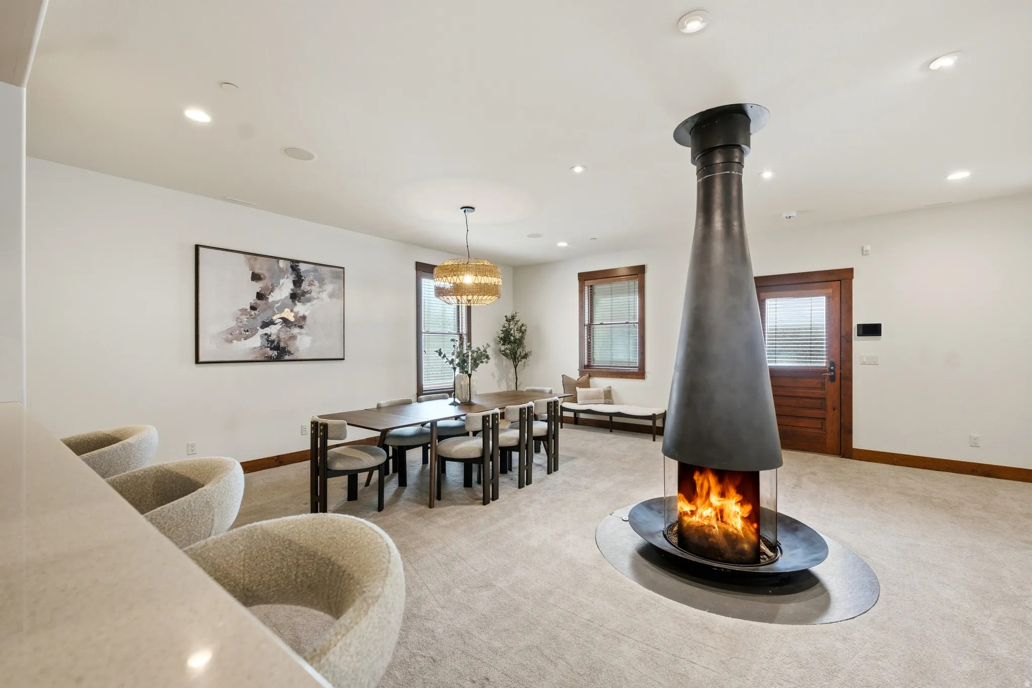 Central freestanding fireplace, recessed ceiling lighting, wood-trimmed windows, wood door with integrated blinds, and plush carpet flooring