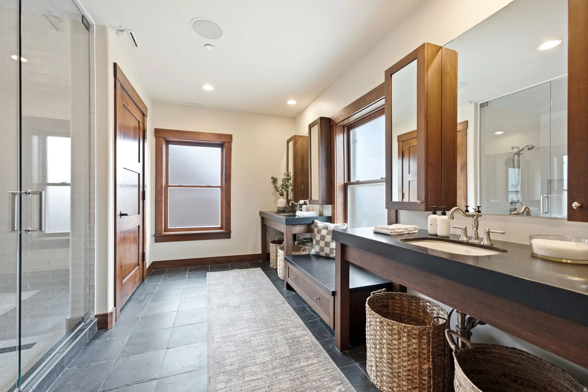Spa-inspired primary bathroom featuring a dual-sink vanity with dark wood cabinetry and black countertop, framed mirrors with integrated lighting, a walk-in shower with frameless glass enclosure, slate-finish tile flooring, and abundant natural light from