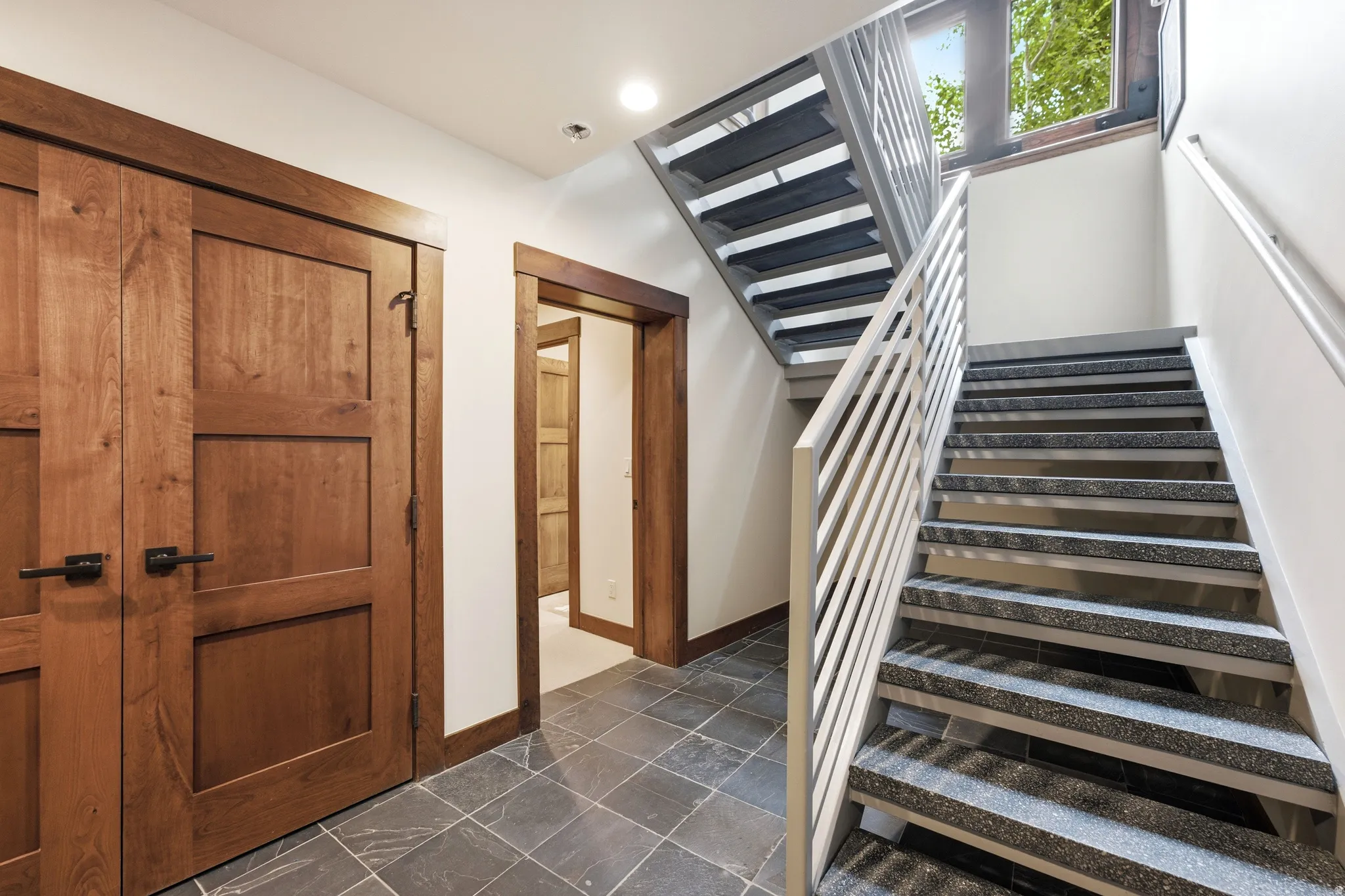 Wood doors with dark hardware, square tile flooring, modern staircase with metal railings, and natural light from transom windows