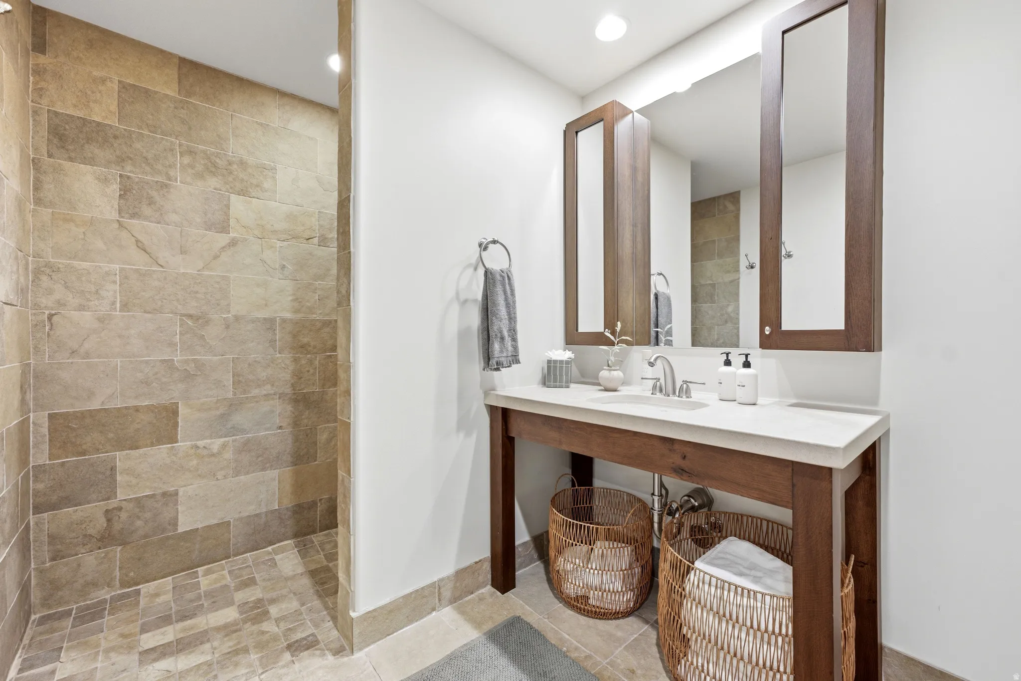 Walk-in shower with natural stone tile, wood-finish vanity, integrated sink, and recessed lighting