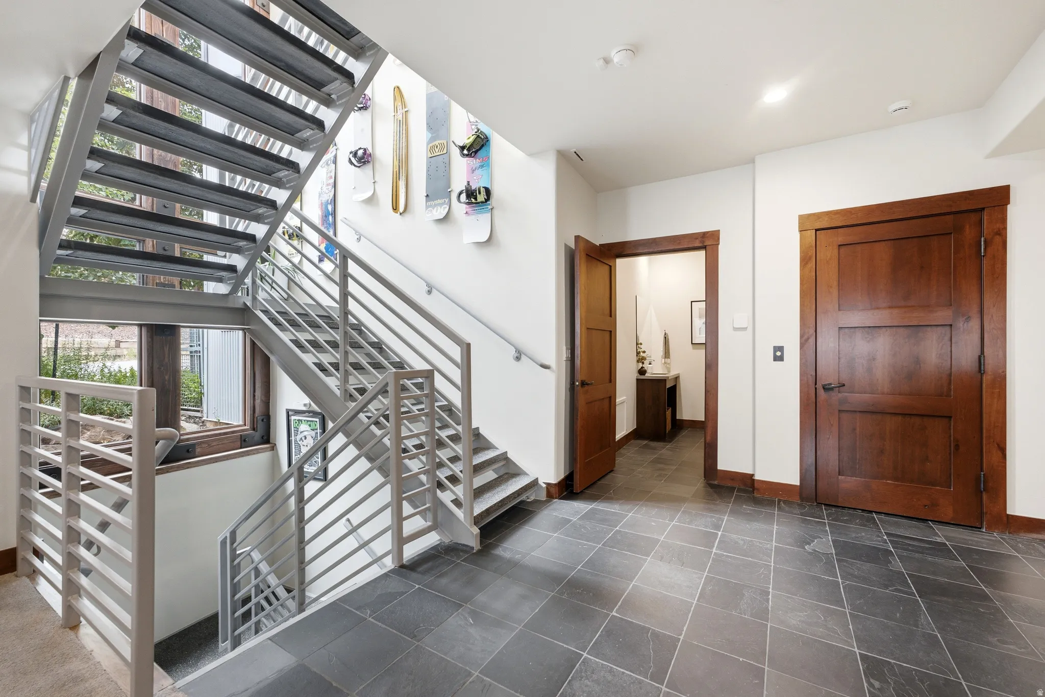Open-riser metal staircase with horizontal railings, dark wood-finish doors, dark wood window trim, and dark tile flooring