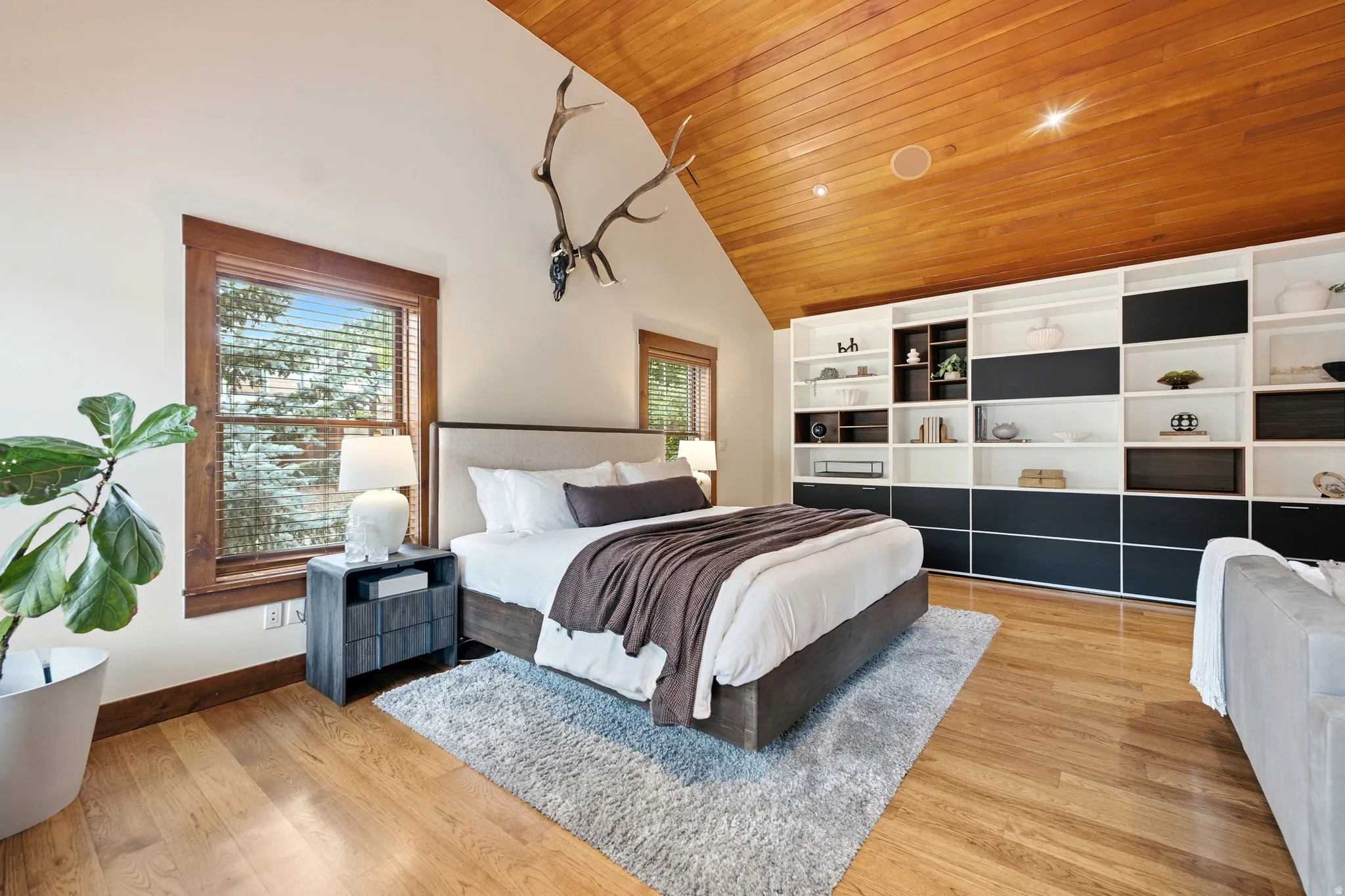 Vaulted wood-paneled ceiling, integrated wall shelving with dark-fronted storage, wood-finish flooring, and large windows with wood trim