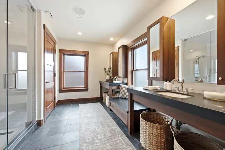 Spa-inspired primary bathroom featuring a dual-sink vanity with dark wood cabinetry and black countertop, framed mirrors with integrated lighting, a walk-in shower with frameless glass enclosure, slate-finish tile flooring, and abundant natural light from