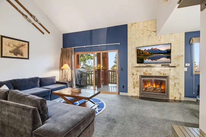 Vaulted ceiling, fireplace with natural stone surround, wall-mounted television, sliding glass door, and exterior balcony