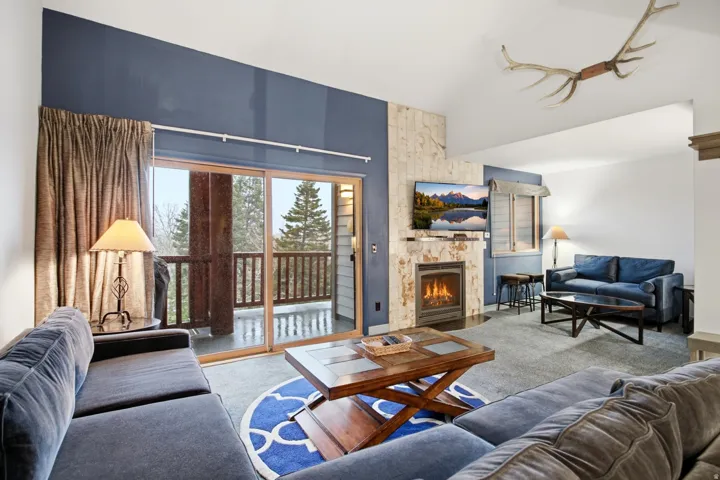 Vaulted ceiling with an accent wall, stone-clad fireplace, and sliding glass doors opening to a balcony with wood railings