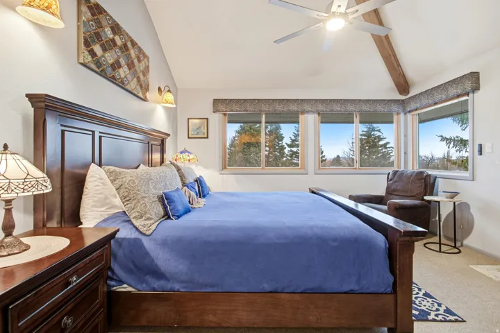 Vaulted ceiling with exposed wood beam, multiple windows, ceiling fan with integrated lighting, wall-mounted sconces, and carpeted flooring