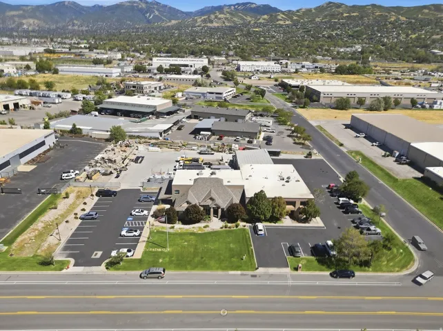 Commercial property featuring multiple buildings, expansive paved parking areas, a landscaped green space, and a mountainous backdrop