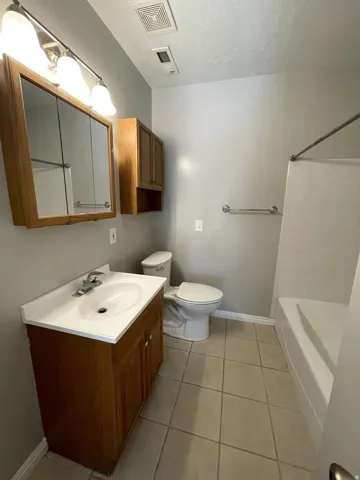 Bathroom featuring a wood-finish vanity with an integrated sink, coordinating wood-finish medicine cabinet, a toilet, a bathtub, and neutral tile flooring