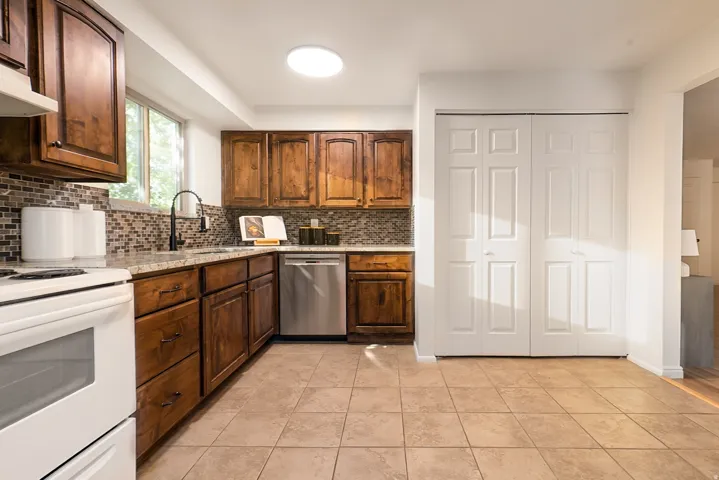 Wood-finish cabinetry with dark hardware, mosaic tile backsplash, granite-style countertops, stainless steel dishwasher, and ceramic tile flooring