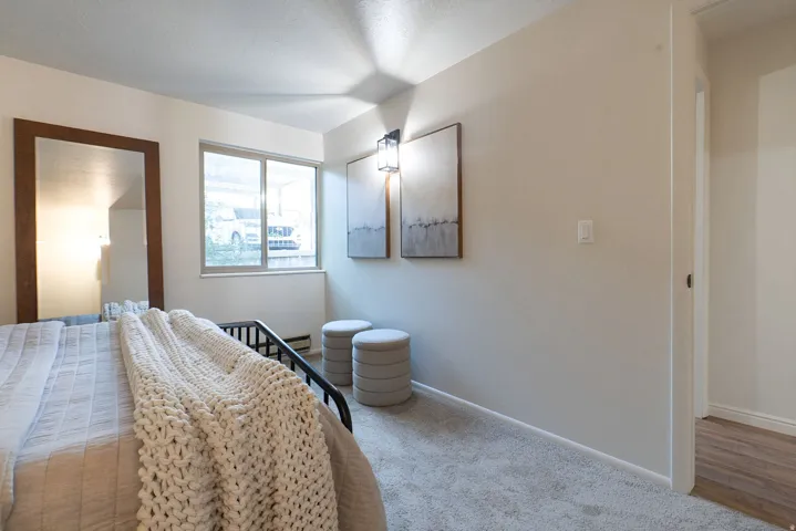 Neutral-toned wall paint, light-colored carpeting, a window with a neutral frame, a wall-mounted light fixture, and a doorway with wood-finish flooring visible beyond