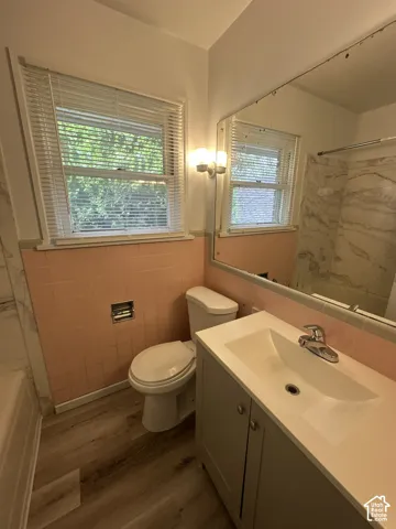 Bathroom featuring a single window with blinds, a wall-mounted light fixture, a vanity with an integrated sink, a toilet, and wood-finish flooring