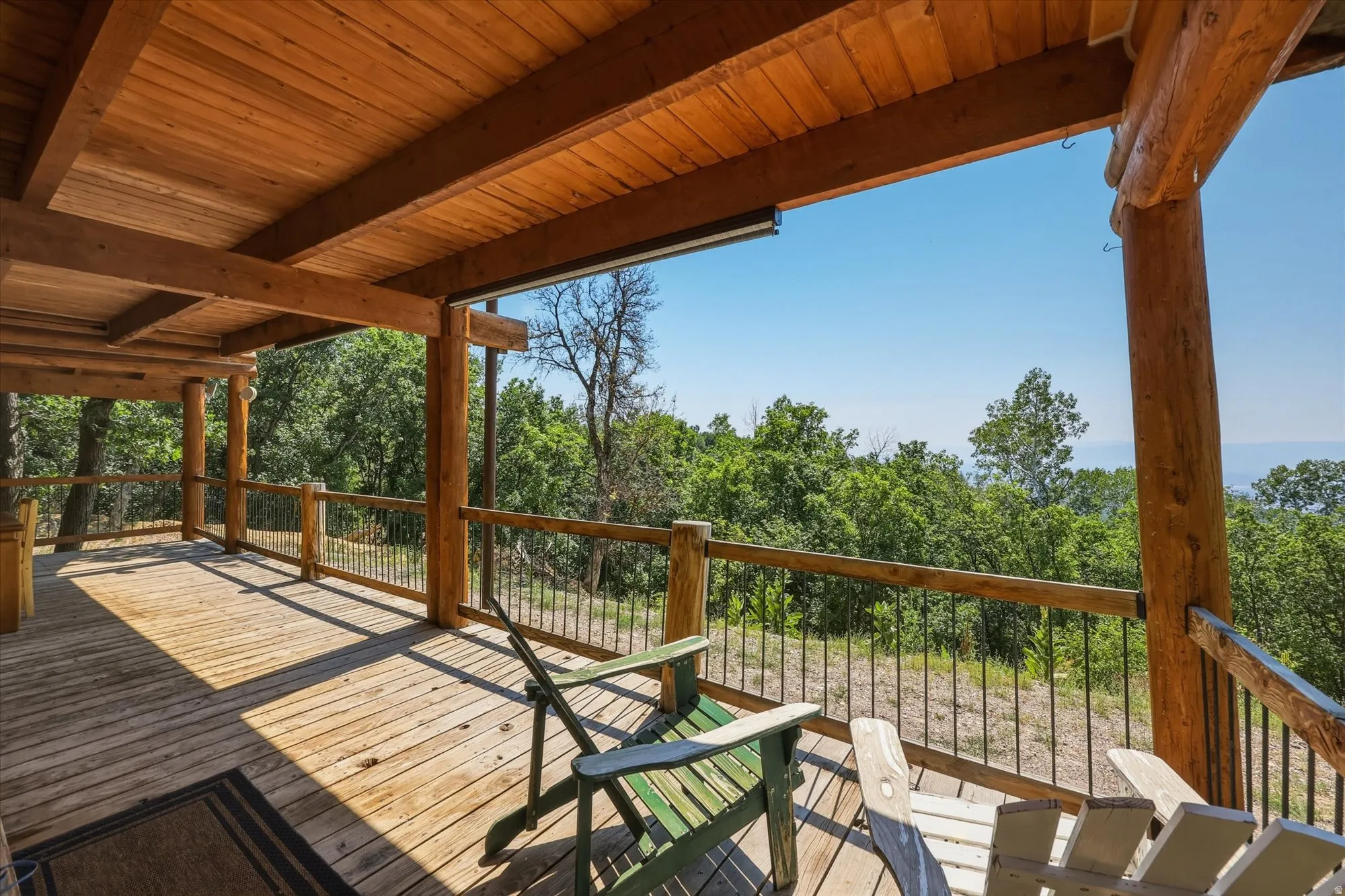 Expansive outdoor deck featuring wood flooring, wood beam ceiling, wood railing with metal balusters, and a natural tree-lined backdrop