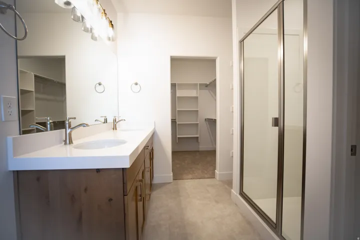 Full primary bath with stained wood cabinets, dual sinks, quartz slab countertops, large walk-in shower and spacious walk-in closet as part of primary suite.