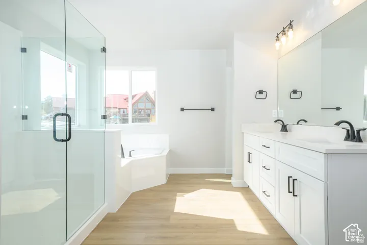 Dual vanity with white cabinetry and countertop, glass-enclosed shower, corner soaking tub, and wood-finish flooring