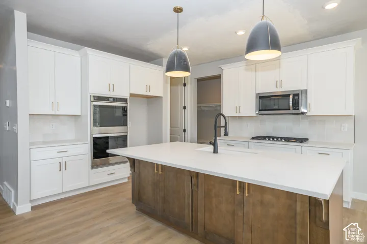 Kitchen featuring a central island with a sink, white cabinetry, integrated double ovens, a built-in microwave, and wood-finish flooring