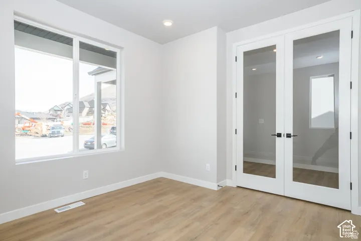 Light wood-finish flooring, white walls, recessed ceiling lighting, a large window, and glass French doors with matte black hardware