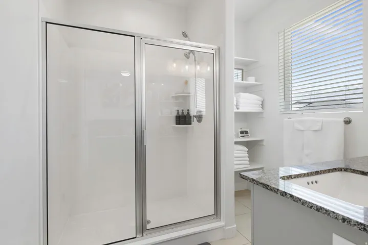 Glass-enclosed shower, built-in shelving, window with blinds, and a built-in sink with stone-finish countertop